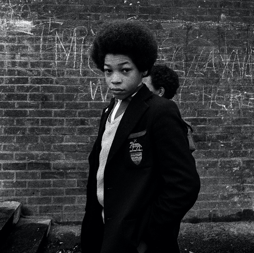 Young boy looking directly at the camera and wearing a school uniform. He is stood in front of a brick wall with graffiti on.