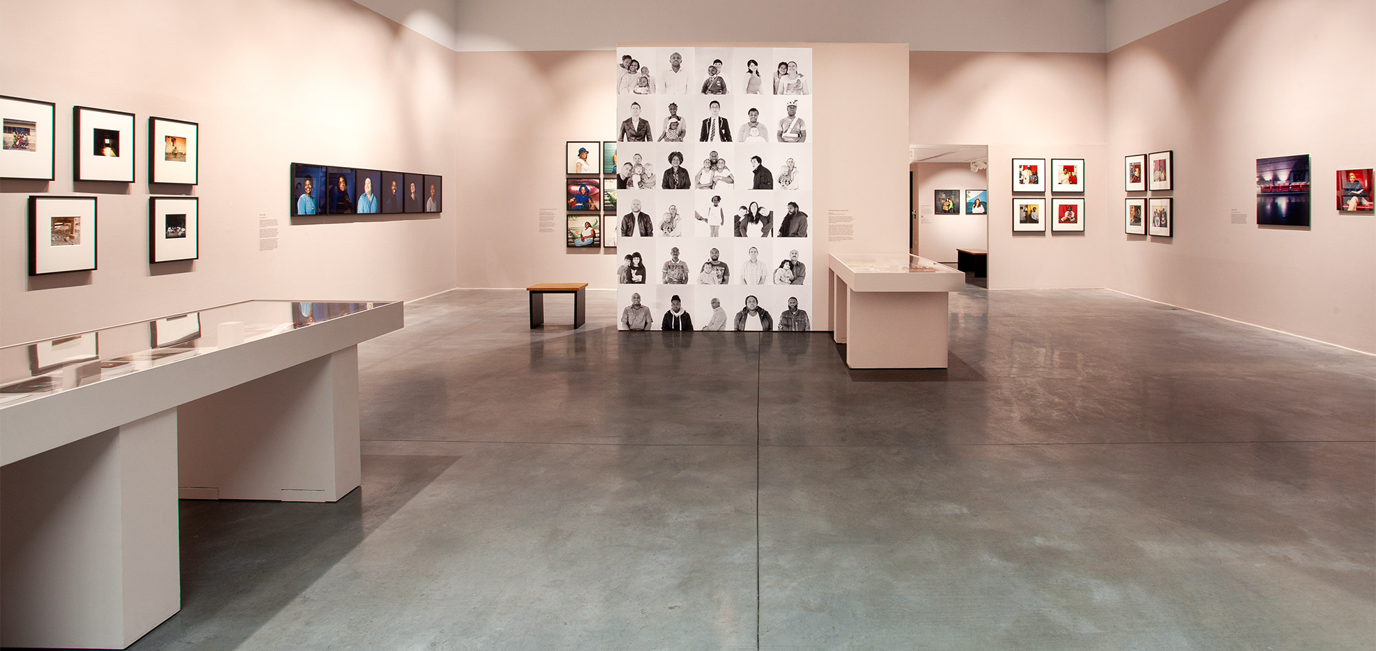 Modern art gallery with framed photographs on soft pink walls, a large black-and-white portrait display in the centre, and glass-topped display tables