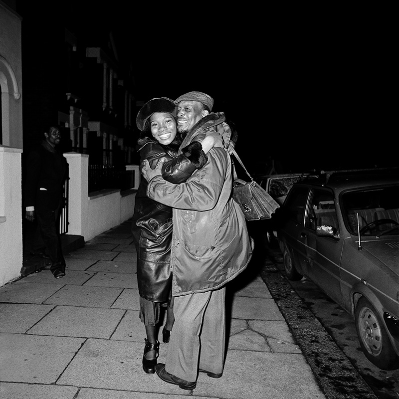 Two smiling people hug on a street at night next to a car. A passerby walks behind them.