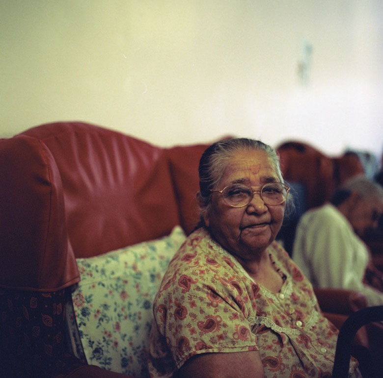 The residents pass their days in this room. Resident sits in the living room in a line of red armchairs, by  Sonal Kantaria, c. 2012. 