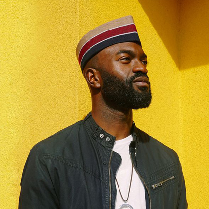 Photograph of Inua Ellams by Caleb Femi. Ellams looks towards the right hand corner of the image, appearing to be gazing in to the distance. He is bearded and against a bright yellow wall.