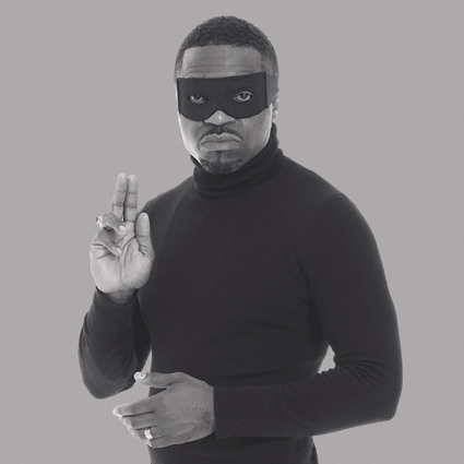 A black and white photograph of the artist Faisal Abdu'Allah dressed in a black poloneck. He has on an apparent superhero eye mask and is holding up two of his fingers on his right hand in an almost religious pose. His left hand is crossed gently across his body.  