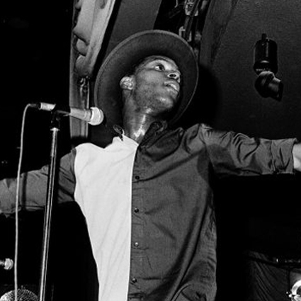 A black and white photograph of a man on stage performing in to a microphone. His hat is tilted upwards and his shirt is split in to two different colours. Sweat is visible on his neck from the exertion of performing. 