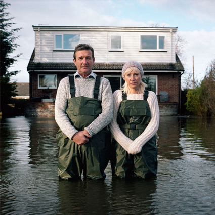 Two people look outwards while standing up to their knees in what looks like a lake. Their house behind them is also partially submerged in water. They are wearing matching green overalls. Photograph from Gideon Mendel's Drowning World