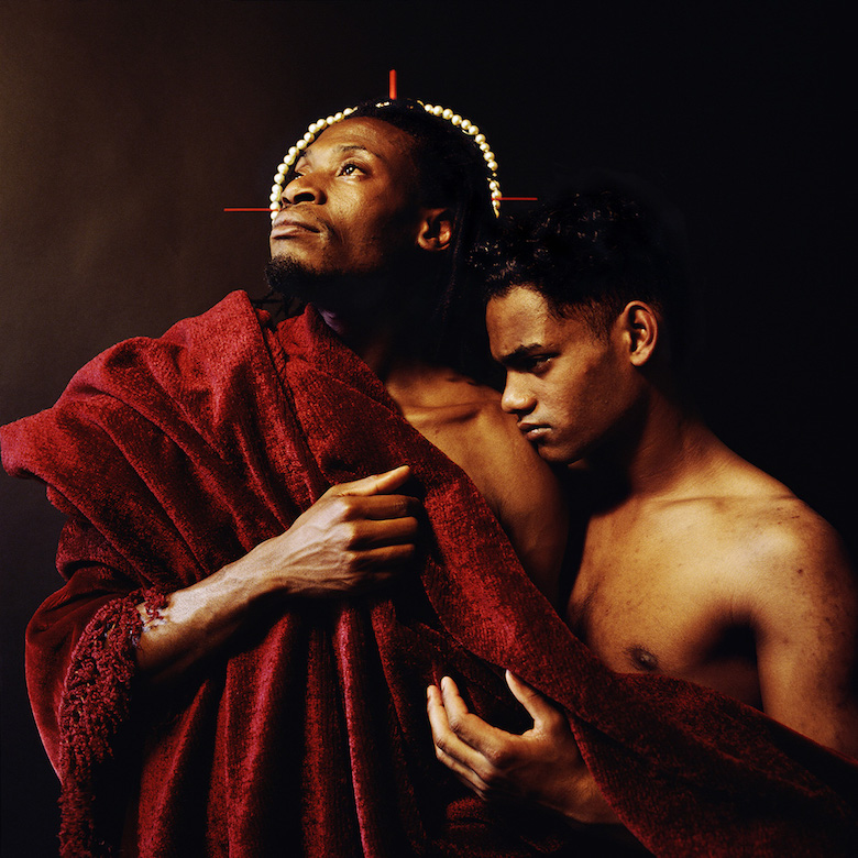Two men wrapped in a red blanket. One is wearing a pearl crown and looking upward, while the other rests his head on the firsts shoulder.