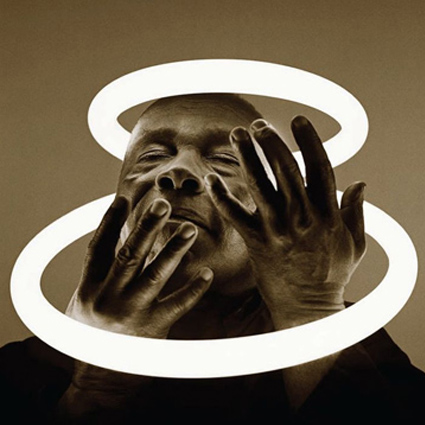 Sepia-toned photograph of a man with his hands up around his face. He is surrounded by two halo-like glowing circles and has his eyes half closed as if he were bathing in the light they provide.