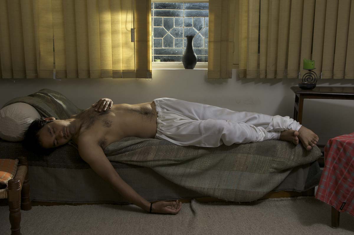 Man laid on a bed with his eyes closed and arm draped on the floor. There is a window behind him with yellow curtains.