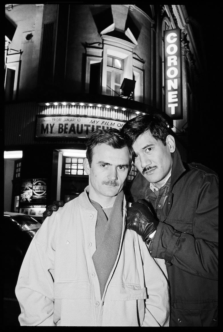 Two men stood in from of a cinema, with large bright lights. They both have dark hair and a mustache.