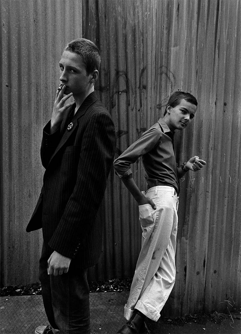 Two young men staring directly at the camera. One is smoking and the other leant against a wall with hand in pocket.
