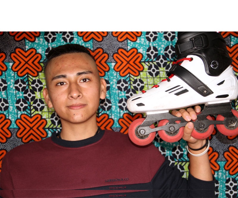 A young person holding a white roller blade looks directly at the camera. There is a colourful patterned backdrop behind them.